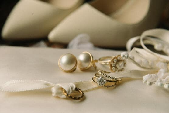 Close-up of pearl earrings, gold rings with diamonds, and a ribbon with rings on a cream-colored surface; blurred white high-heeled shoes in the background. by Justin Salem Meyer