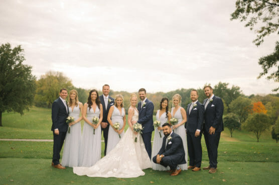 A wedding party poses outdoors on a golf course, with six women in light gray dresses and five men in dark suits surrounding a bride and groom, all smiling at the camera. Lush green trees and cloudy skies are in the background. by Justin Salem Meyer