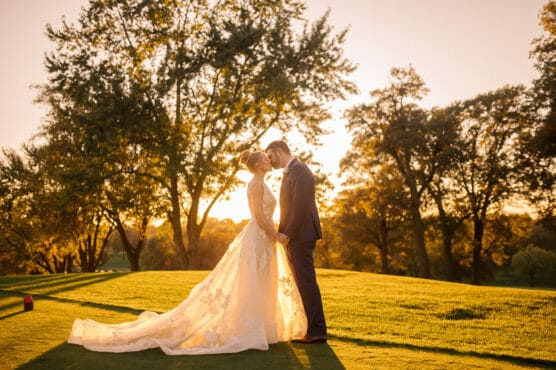 A bride and groom stand close together, holding hands and touching foreheads, on a sunlit grassy field with trees in the background during golden hour. The bride wears a long, flowing gown and the groom is in a suit. by Justin Salem Meyer