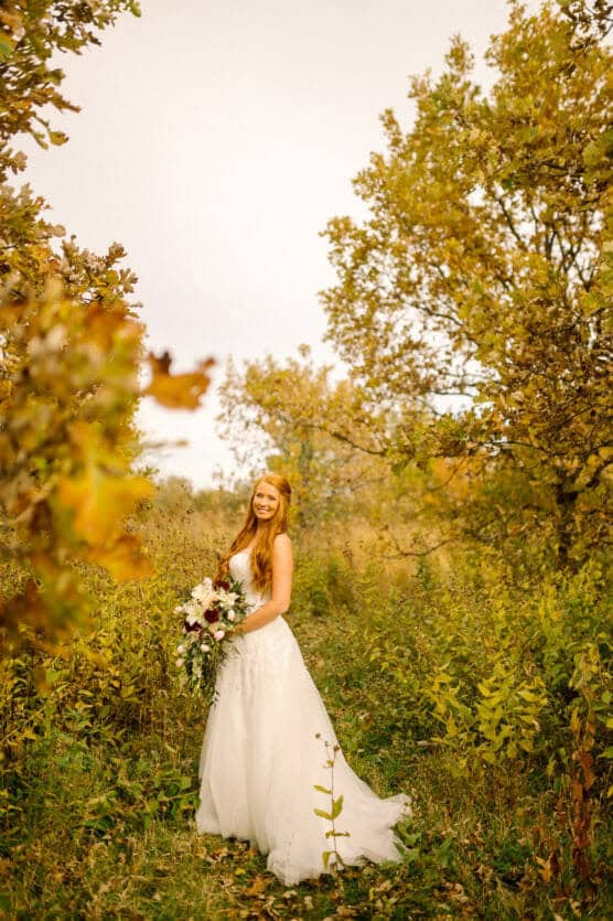 A bride in a white dress stands outdoors among tall green and yellow foliage, holding a bouquet of flowers and smiling at the camera on a bright, sunny day. by Justin Salem Meyer
