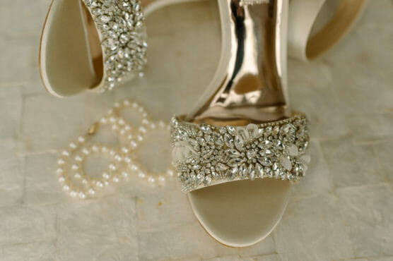 A close-up of an elegant high-heeled sandal with sparkling rhinestone embellishments and a pearl necklace arranged beside it on a textured, light-colored surface. by Justin Salem Meyer