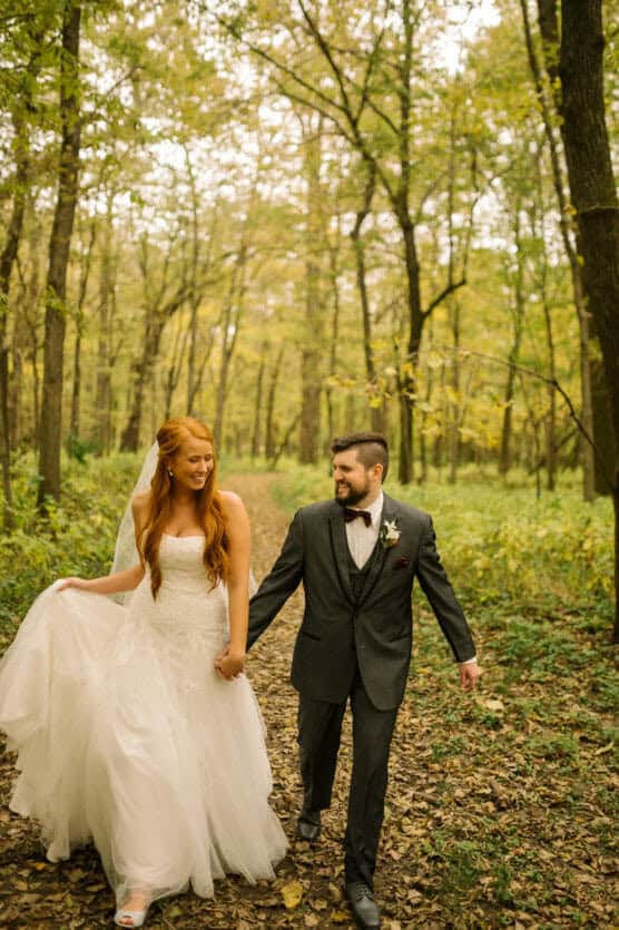 A bride in a white gown and a groom in a dark suit walk hand in hand along a forest path, smiling at each other, surrounded by tall trees with green and yellow leaves. by Justin Salem Meyer