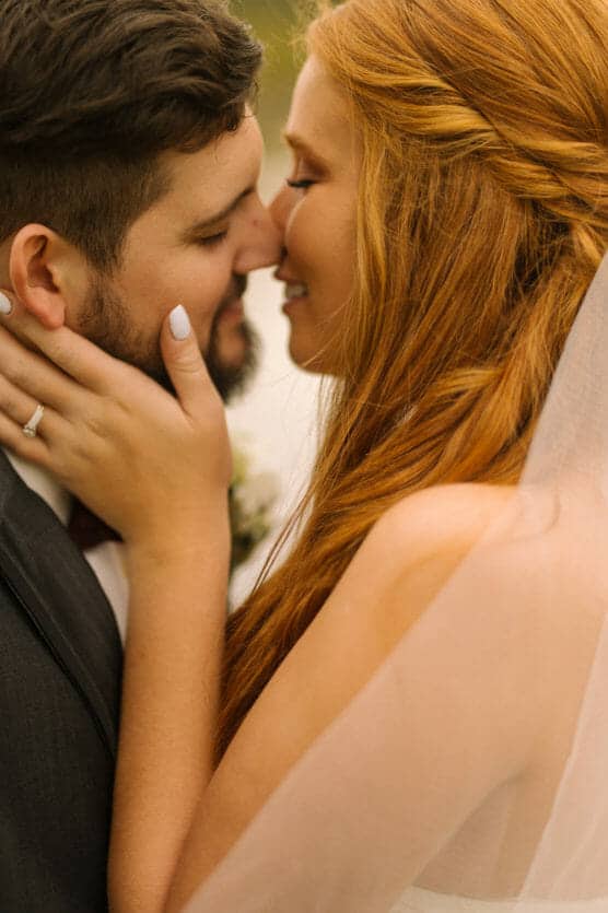 A close-up of a bride and groom about to kiss outdoors. The bride, with long red hair and a veil, gently holds the groom’s face, showing her engagement ring, while both smile softly with closed eyes. by Justin Salem Meyer
