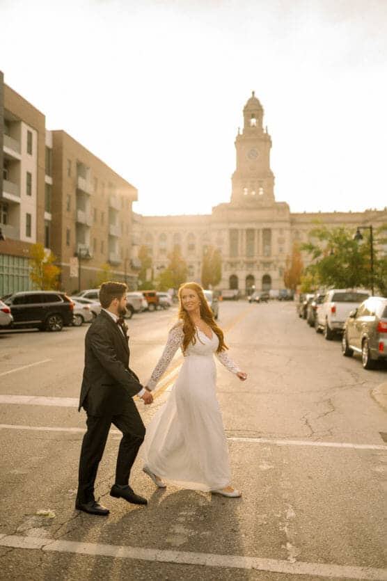 A bride and groom walk hand-in-hand across a city street at sunset. The bride wears a long white dress and the groom is in a black suit. Cars are parked along the road, and a historic building stands in the background. by Justin Salem Meyer