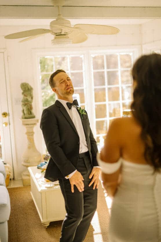 A man in a black tuxedo stands indoors, making a playful face and gesture, while a woman in an off-the-shoulder white dress stands in the foreground with her back to the camera. Sunlight streams through large windows behind them. by Justin Salem Meyer