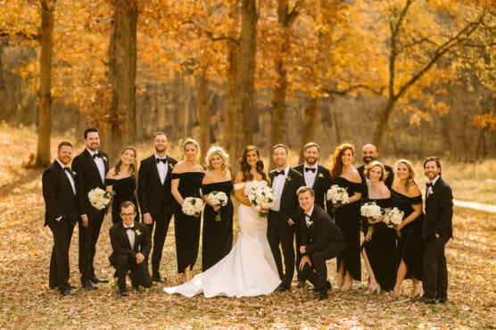 A wedding party poses outdoors among autumn trees. The bride in a white gown stands at the center, surrounded by bridesmaids in black dresses and groomsmen in black suits with bow ties. Everyone is smiling and holding flowers. by Justin Salem Meyer