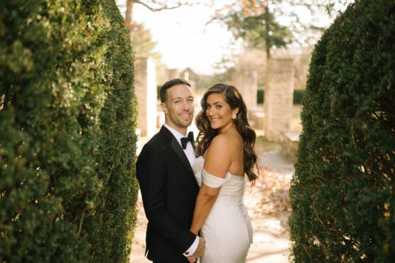 A bride in an off-shoulder white dress and a groom in a black tuxedo stand close together outdoors, smiling and holding hands between two tall green hedges. by Justin Salem Meyer