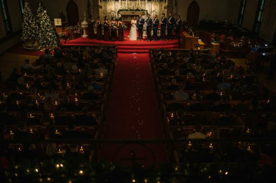 A wedding ceremony taking place in a dimly lit church, with the bride, groom, and wedding party at the altar. Guests are seated in pews, holding candles, and a decorated Christmas tree is visible on the left. by Justin Salem Meyer