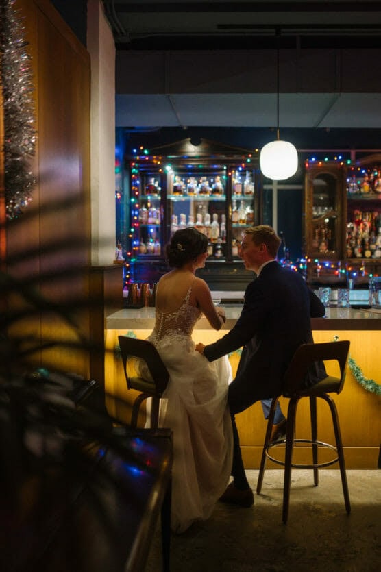 A bride and groom sit together at a bar decorated with colorful string lights. The bride wears a white dress with lace details, and the groom is in a suit. They hold hands and smile at each other in a cozy, festive setting. by Justin Salem Meyer