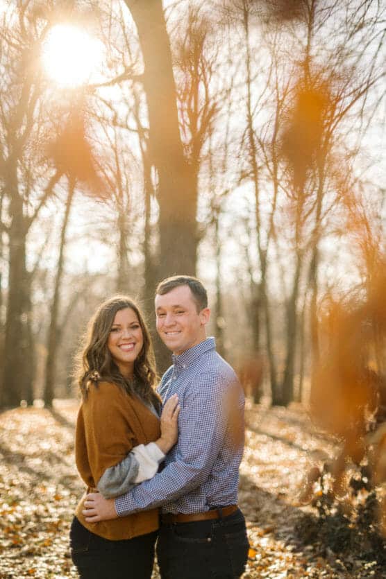 A couple stands close together, smiling at the camera in a sunlit forest with bare trees and fallen leaves. Warm sunlight shines through the branches, creating a golden, autumnal atmosphere. by Justin Salem Meyer