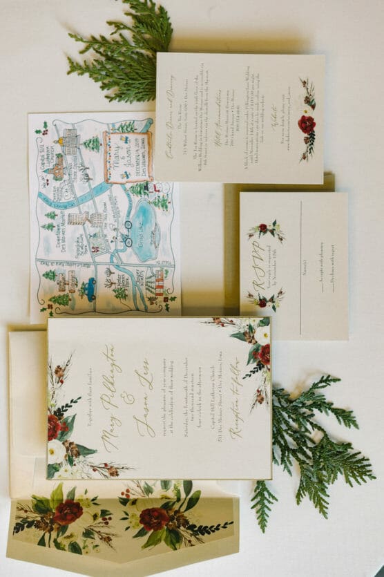A floral-themed wedding invitation suite is displayed, including an illustrated map, RSVP card, and invitation envelope, all decorated with botanical accents. Greenery sprigs are arranged around the stationery. by Justin Salem Meyer
