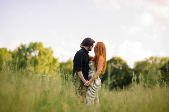 A couple stands close together in a grassy field, holding hands and smiling at each other. The woman has long red hair and wears a striped dress; the man has dark hair and wears a black shirt. Trees and sky are in the background. by Justin Salem Meyer