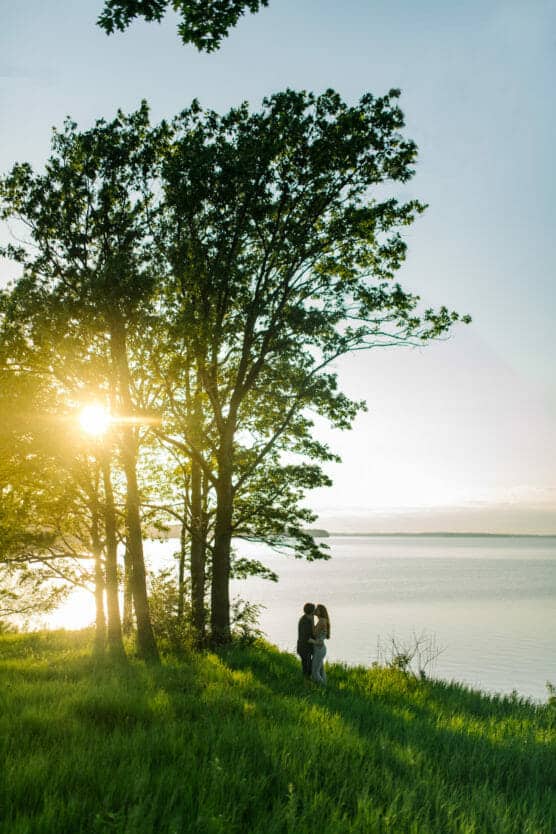 A couple stands close together on a grassy hillside by a lake, framed by tall trees. The sun shines through the leaves, creating a serene and romantic atmosphere near the water’s edge. by Justin Salem Meyer