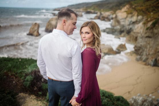 A couple stands holding hands on a cliff overlooking a sandy beach and rocky coastline. The man wears a white shirt and blue pants, while the woman in a maroon dress looks back at the camera. by Justin Salem Meyer