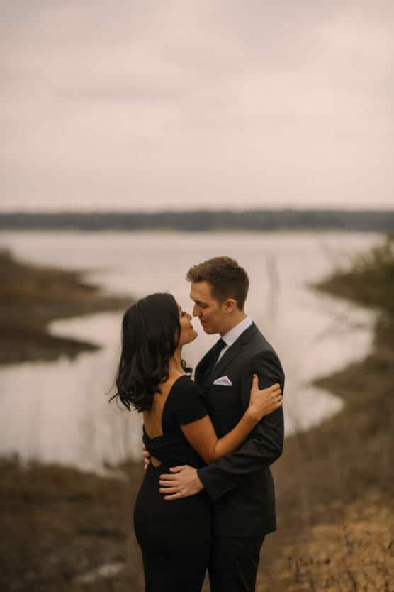 A couple dressed formally embraces and gazes at each other by a calm riverside on an overcast day, with muted natural scenery in the background. by Justin Salem Meyer