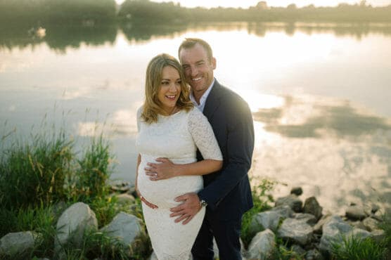 A smiling couple stands by a lake at sunset. The woman, wearing a white lace dress, is visibly pregnant, and the man in a suit embraces her from behind, both looking happy and relaxed by the water’s edge. by Justin Salem Meyer