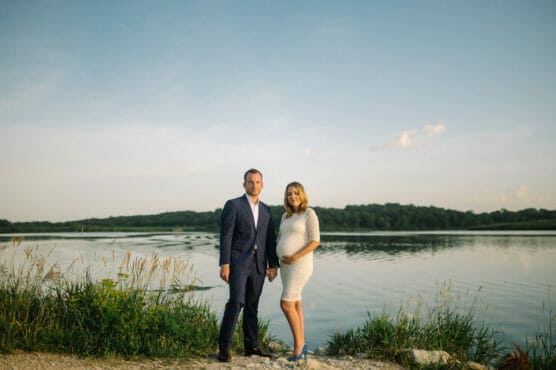 A man in a suit and a pregnant woman in a white dress stand together by a calm lake, surrounded by tall grass and greenery, under a clear blue sky. by Justin Salem Meyer