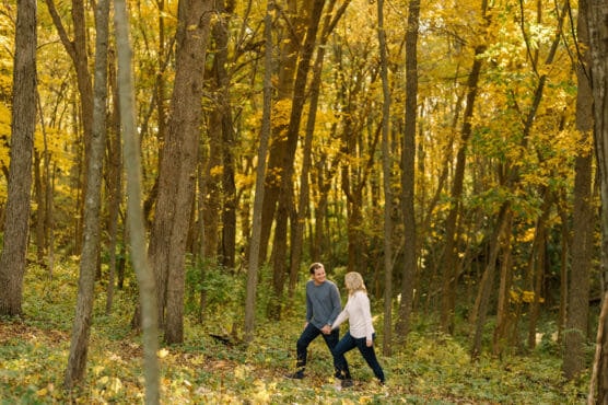 A couple holds hands and walks together through a sunlit forest with tall trees and golden autumn leaves, surrounded by green undergrowth. by Justin Salem Meyer