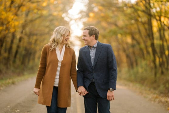 A couple walks hand in hand down a tree-lined road during autumn, smiling at each other. The trees have golden leaves and sunlight filters through the branches, creating a warm, romantic atmosphere. by Justin Salem Meyer