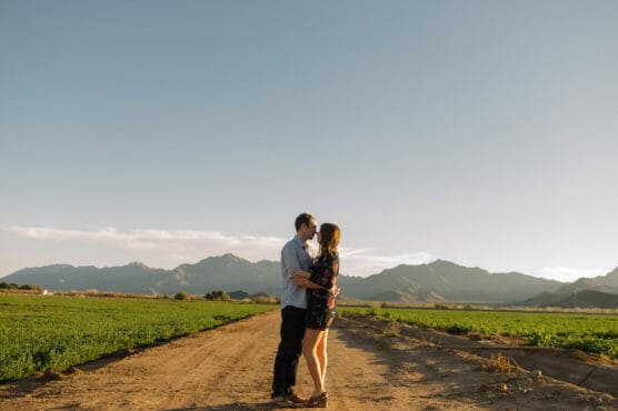 A couple stands facing each other on a dirt road surrounded by green fields, embracing under a clear sky with mountains in the background. The scene is bathed in warm sunlight. by Justin Salem Meyer