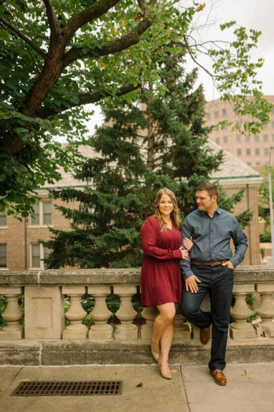 A woman in a burgundy dress and a man in a blue shirt and dark jeans stand together on a stone terrace, arm in arm, smiling with trees and a brick building in the background. by Justin Salem Meyer