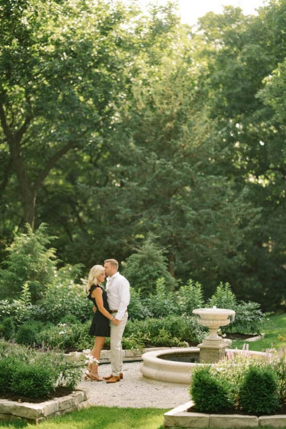 A couple stands close together in a lush, sunlit garden near a round fountain, surrounded by greenery and trees, sharing an intimate moment. by Justin Salem Meyer