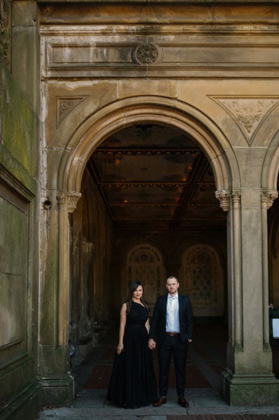 A woman in a black dress and a man in a suit stand hand in hand beneath an ornate stone archway, posing for a photo in a grand, historic building. by Justin Salem Meyer