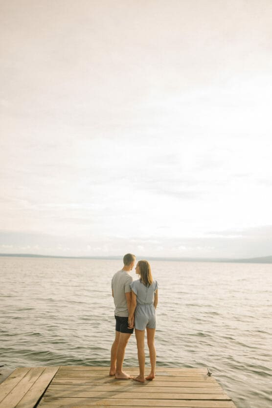 A couple stands barefoot on a wooden dock, holding hands and gazing out over a calm, expansive body of water under a bright, lightly clouded sky. by Justin Salem Meyer
