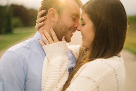 A couple embraces outdoors at sunset, smiling with their faces close together. The woman gently holds the mans face, showing an engagement ring, and both look happy and affectionate in the warm light. by Justin Salem Meyer