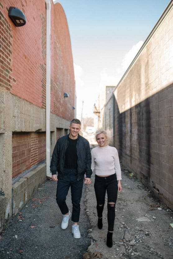 A smiling couple walks hand-in-hand down an urban alleyway lined with brick and concrete walls on a sunny day. Both wear casual dark clothing and look happy together. by Justin Salem Meyer