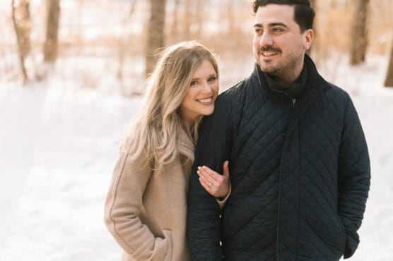 A woman in a tan coat smiles while holding the arm of a man in a dark quilted jacket. They stand close together outdoors in a snowy, sunlit forest. by Justin Salem Meyer