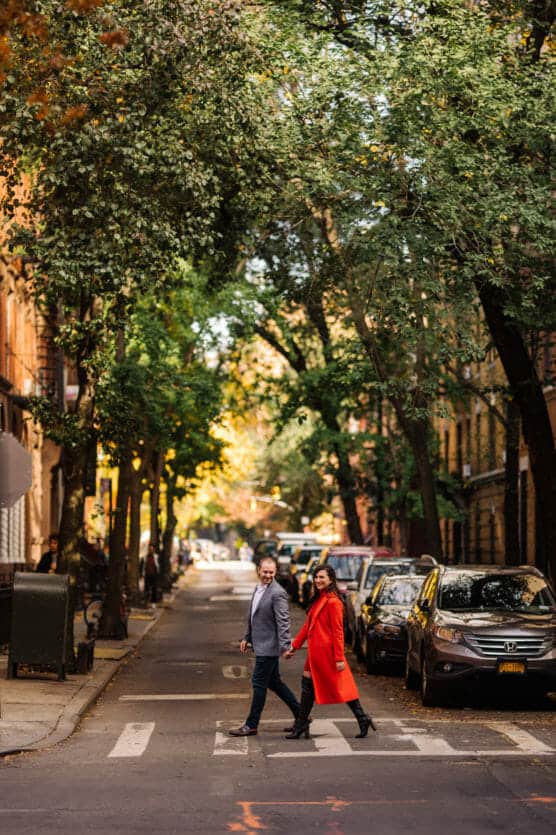 A man and woman hold hands while crossing a tree-lined street. The woman wears a bright red coat and the man wears a gray blazer. Parked cars and brownstone buildings line the road, with sunlight filtering through the trees. by Justin Salem Meyer