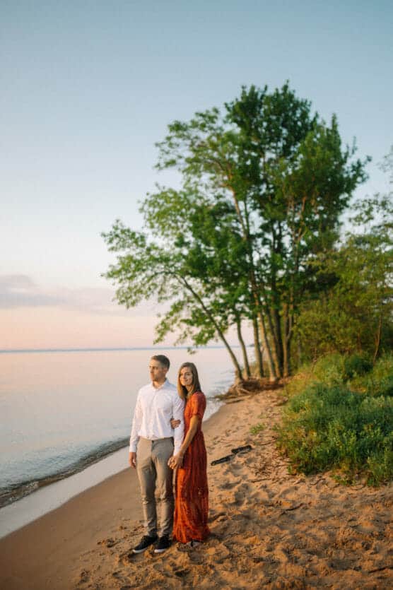 A couple stands hand in hand on a sandy beach at sunset. The man wears a white shirt and gray pants, while the woman wears a long rust-colored dress. Green trees and calm water are in the background under a clear sky. by Justin Salem Meyer
