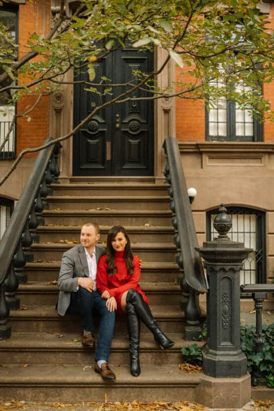 A man in a gray blazer and a woman in a red dress sit together on brownstone steps outside a brick building, surrounded by autumn leaves and greenery. by Justin Salem Meyer