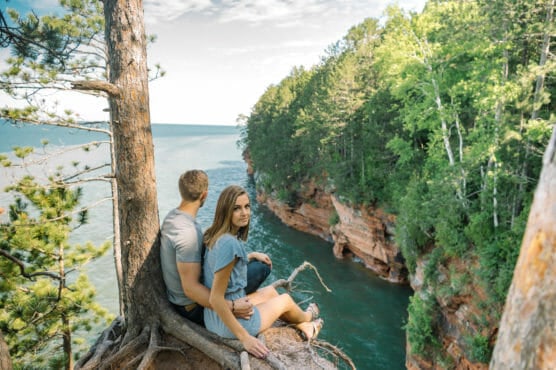 A couple sits on tree roots at the edge of a cliff overlooking blue water and forested red rock cliffs, with pine trees and lush greenery surrounding them under a partly cloudy sky. by Justin Salem Meyer