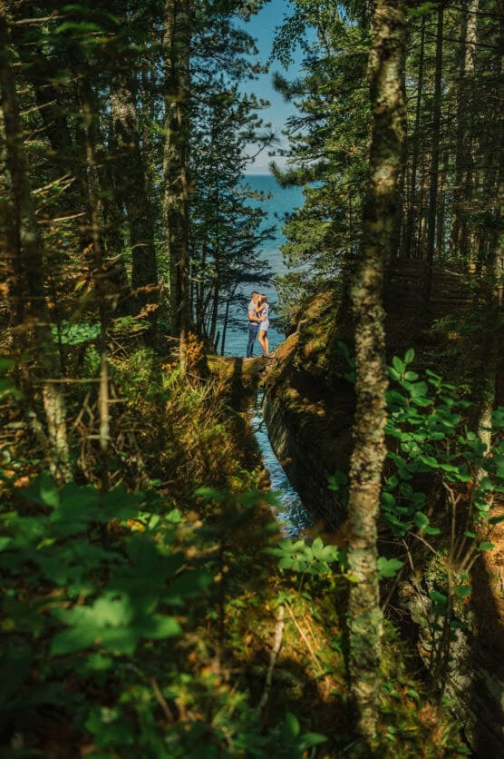 A couple embraces in a sunlit forest clearing near a narrow stream, with tall trees surrounding them and a view of a blue lake visible through the trees in the background. by Justin Salem Meyer
