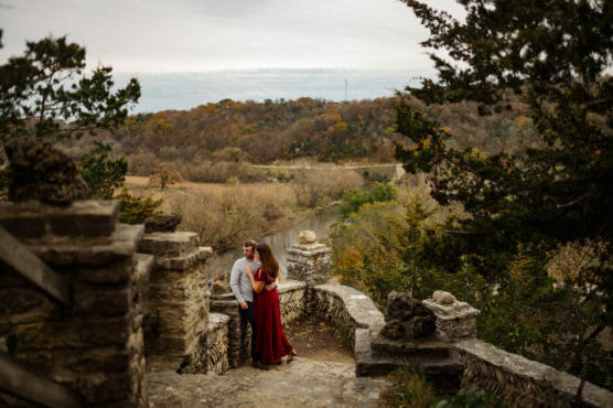 A couple stands close together on a stone pathway overlooking a scenic, tree-covered valley and river under a cloudy sky, surrounded by old stone walls and autumn foliage. by Justin Salem Meyer