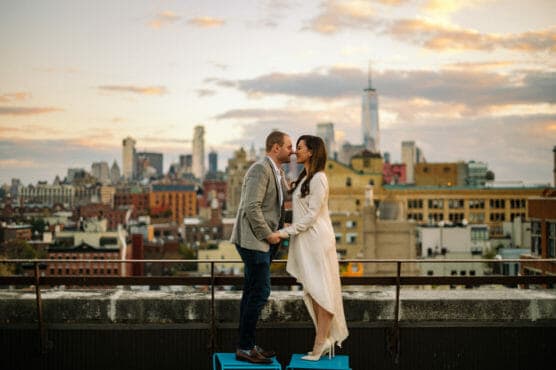 A couple stands on a rooftop at sunset, holding hands and about to kiss, with the city skyline and tall buildings in the background. by Justin Salem Meyer