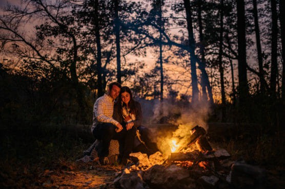 A couple sits close together by a campfire in a forest at dusk. Warm firelight illuminates their faces as trees and a colorful evening sky create a peaceful, intimate atmosphere in the background. by Justin Salem Meyer