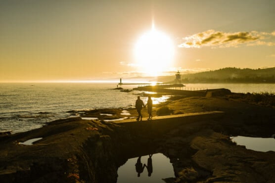 Two people walk along a rocky shoreline at sunset, with the sun low in the sky, creating bright reflections on the water and wet rocks. Distant lighthouses and hills are visible under a golden sky. by Justin Salem Meyer
