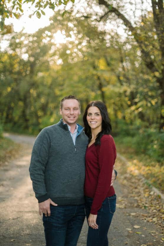 A man and woman stand close together on a wooded path, smiling at the camera. Sunlight filters through the trees, and fallen leaves cover parts of the ground. Both are dressed in casual autumn clothing. by Justin Salem Meyer