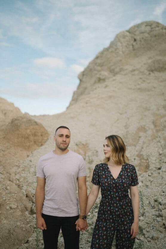 A man and woman stand holding hands in front of rocky, sandy hills under a blue sky. The man wears a light t-shirt and dark pants; the woman wears a patterned jumpsuit and looks at him while he looks ahead. by Justin Salem Meyer