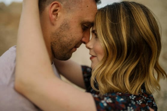 A close-up of a couple embracing, facing each other with their foreheads touching, eyes closed, and arms around each other, sharing an intimate and affectionate moment outdoors. by Justin Salem Meyer