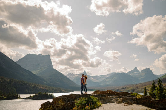 A couple stands embracing on a rocky ledge overlooking a scenic landscape of mountains, a river, and forests under a partly cloudy sky. by Justin Salem Meyer