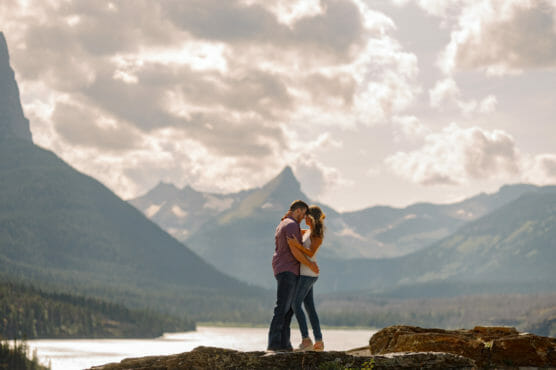 A couple embraces on a rocky ledge with mountains, a river, and a partly cloudy sky in the background, creating a romantic and scenic outdoor setting. by Justin Salem Meyer