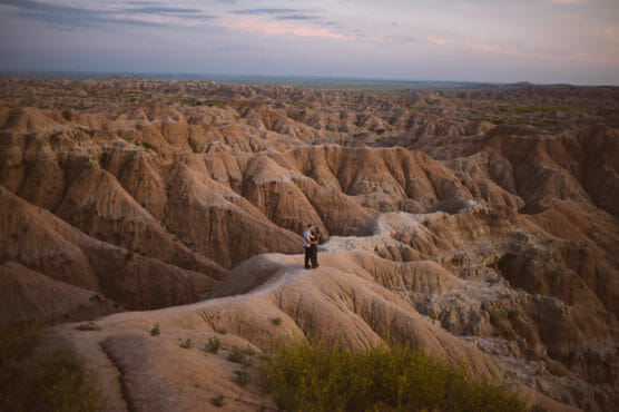 A couple embraces while standing on a rugged, rocky ridge in a vast, eroded landscape at sunset, surrounded by dramatic, layered formations under a partly cloudy sky. by Justin Salem Meyer
