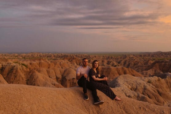 A couple sits on rocky terrain at sunset, surrounded by dramatic, rugged formations. The sky is partly cloudy with a warm, golden light illuminating the landscape and the pair, who gaze calmly into the distance. by Justin Salem Meyer