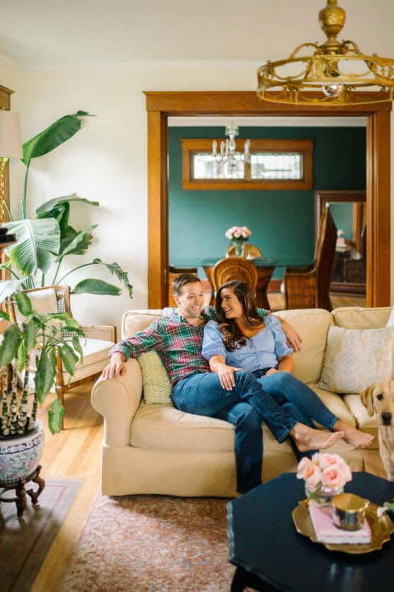 A couple sits closely together, smiling on a cream-colored sofa in a cozy, well-lit living room with plants and a glimpse of a dining room in the background. A dog is partially visible in the lower right corner. by Justin Salem Meyer