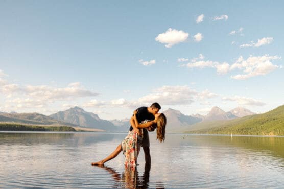 A couple stands in shallow water with mountains in the background, as the man dips the woman in a romantic pose under a blue sky with scattered clouds. by Justin Salem Meyer