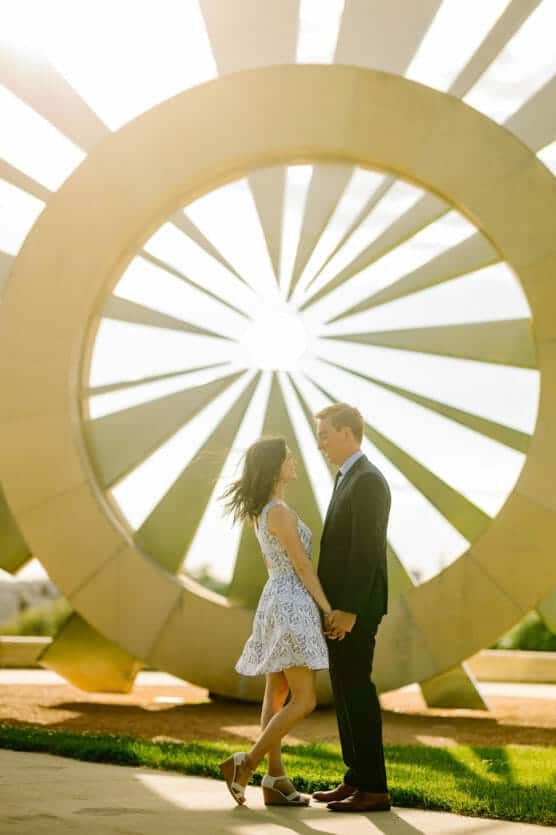 A couple stands holding hands, facing each other and smiling, in front of a large, circular, sunburst sculpture. The sunlight creates a warm glow around them, highlighting their joyful expressions. by Justin Salem Meyer